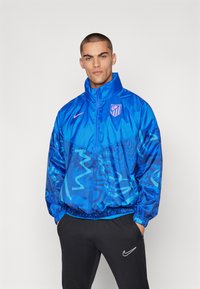 Modello maschile che indossa una giacca mezzo zip a maniche lunghe a motivi blu dell'Atletico Madrid e pantaloni neri Nike, in piedi con le mani nelle tasche contro uno sfondo bianco.