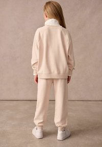 Vaaleanbeige, fleece-vuorattu huppari raglan-hihoilla, yhdistettyinä yhteensopiviin kapeisiin verkkareihin. Valkeat lenkkarit viimeistelevät asukokonaisuuden.