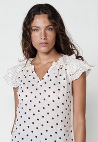 Blusa bianca con pois neri, scollatura a V e maniche a sbuffo con pois. Tessuto leggero, design fluido e vestibilità morbida.