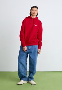 Roter Baumwoll-Hoodie mit einer Fronttasche und Kordelzugkapuze, kombiniert mit blauen weiten Jeans und beigefarbenen Turnschuhen auf einem grünen Boden.