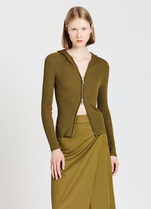Jeune femme aux cheveux blonds courts portant un pull zippé côtelé olive et une jupe enveloppante olive assortie, se tenant devant un fond blanc.