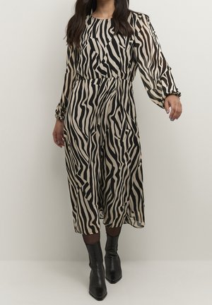 Kobieta ubrana w beżowo-czarną midi sukienkę w paski zebry z długimi rękawami oraz czarne, szpiczaste botki, stojąca na tle jednolitej ściany.