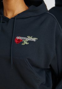 Mørkeblå hettegenser med brodert "Converse"-logo og design av en rød rose, laget av myk bomullsblanding med frontlomme og snorhette.