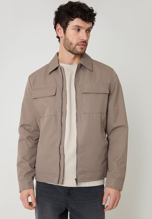 Mand med mørkt krøllet hår iført en beige lynlåstrøje over en lys skjorte og mørke jeans, der kigger til sin venstre side.