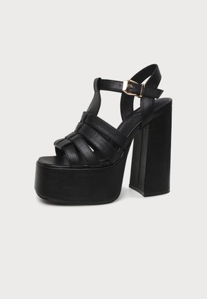 Sandales noires à brides et à plateforme, avec une semelle épaisse, un talon bloc haut et une boucle de cheville dorée, sur un fond uni.