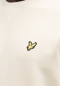 Mikina v krémovej farbe so štruktúrovanou, mäkkou textúrou. Na hrudi výrazné čierno-zlaté vyšívané logo vtáka. Žebrovaný výstrih a švy.