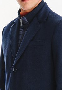 Granatowy płaszcz wełniano-akrylowy z usztywnionym kołnierzem, zapięciem na jeden guzik i zapinaną na zamek wewnętrzną warstwą. Posiada gładką fakturę oraz minimalne detale kieszeni.