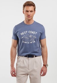 Mėlyna medvilninė marška su balta 'WEST COAST' užrašą, trumpomis rankovėmis ir apvaliu iškirpimu, derinama su smėlio spalvos pritaikytomis kelnėmis.