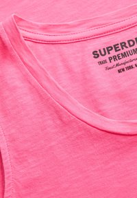 Primer plano de una camiseta sin mangas rosa de algodón que muestra la costura del escote y una etiqueta impresa negra parcial con detalles de la marca y la ubicación.