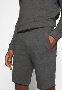 Shorts en coton gris avec des accents rouges, dotés d'une taille élastique à cordon de serrage et de poches latérales. Texture lisse et design décontracté.