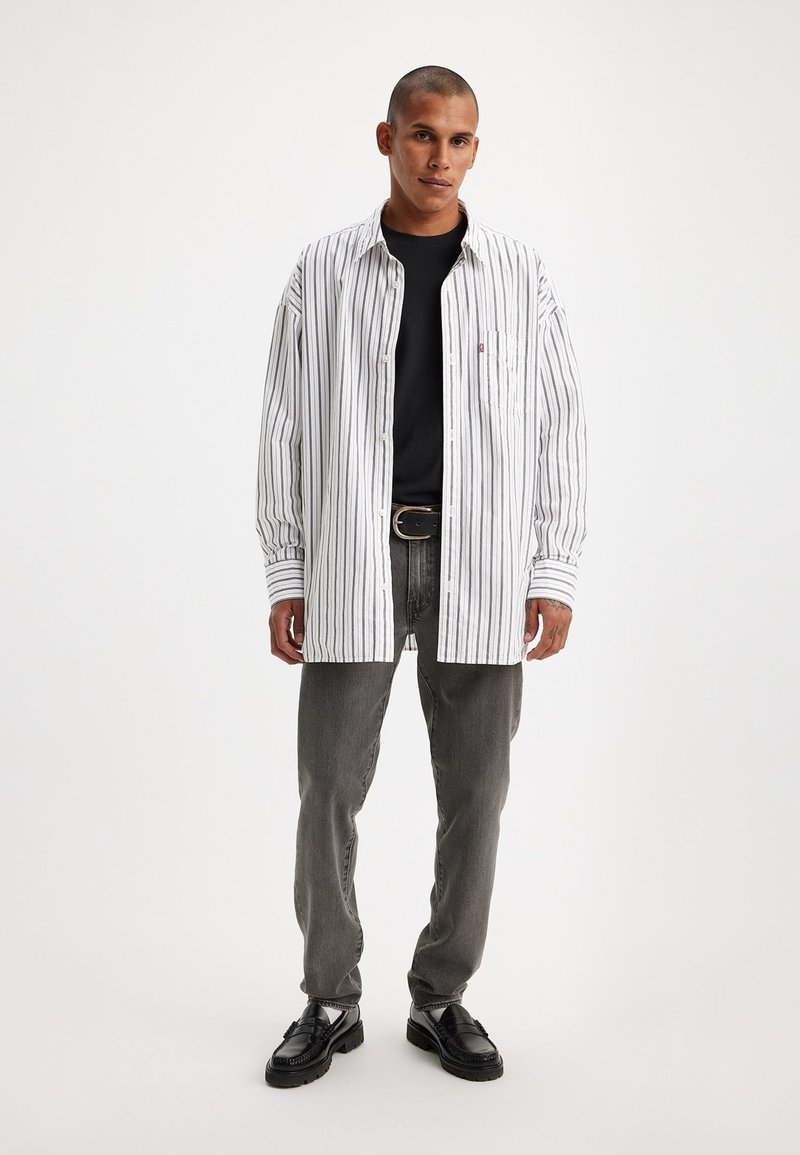 Chemise à rayures à manches longues blanche et noire, associée à un t-shirt noir et un jean gris. Portée avec des chaussures noires, au design classique.