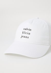 Fehér pamut baseballsapka ívelt karimával. Elöl fekete szöveg olvasható: "calvin klein jeans". Egyszerű dizájn, további díszítések nélkül.
