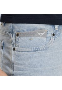 Helleblaue Denim-Jeans mit einer Vordertasche, die einen grauen Stoff mit einem dezenten Logo zeigt. Mit Nähten und schwarzen Hardware-Akzenten.