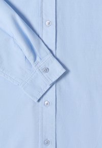 Light blue button-up shirt made of smooth fabric, featuring a pointed collar, long sleeves, and white button accents. Texture is subtly ribbed.