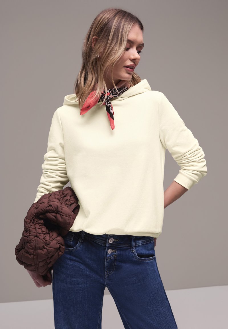 Joven mujer con sudadera con capucha color crema y bufanda roja con estampado, sosteniendo una chaqueta marrón acolchada doblada, de pie frente a un fondo neutro.