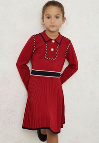 Abito rosso lavorato a maglia con colletto, dettagli con bottoni, rifiniture intrecciate e gonna plissettata con dettagli in contrasto blu navy e beige.