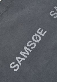 Grå stoff tote bag med teksturert overflate. Har store hvite bokstaver som staver "SAM Sø E." Enkel design uten ekstra detaljer eller mønstre.