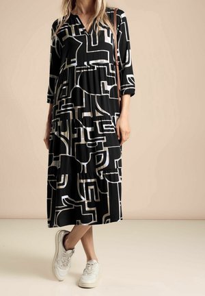 Frau trägt ein schwarzes Midikleid mit weißen abstrakten geometrischen Mustern, dreiviertellangen Ärmeln, weißen Turnschuhen und einer braunen Umhängetasche.