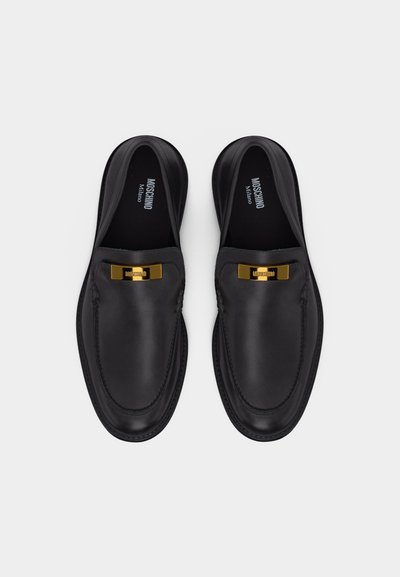Μαύρα δερμάτινα loafers με χρυσές πλακέτες λογότυπου Moschino, στρογγυλεμένες μύτες και εσωτερικές σόλες με το εμπορικό σήμα, απεικονιζόμενα από πάνω σε λευκό φόντο.