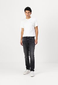 Weißes Baumwoll-T-Shirt mit kleinem Logo auf der Brust, kombiniert mit dunkelgrauen Jeans und weißen Sneakern, stehend vor einem schlichten Hintergrund.