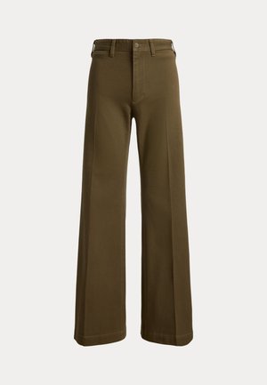 Femme debout les mains dans les poches, portant un cardigan bleu marine sur une chemise bleu clair boutonnée, un pantalon large olive et des chaussures marron.