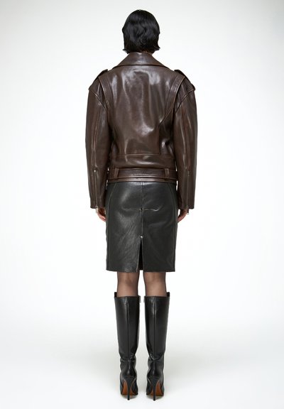 Személy barna bőrdzsekiben, fekete bőrszoknyában, hátul hasítékkal, és térdig érő fekete bőrcsizmában, fehér háttér előtt, háttal állva.