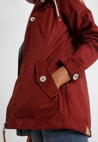 Giacca rossa con finitura testurizzata, dotata di tasche laterali, grandi bottoni, una cerniera e un cappuccio con coulisse. Accentuata da una patch con logo.