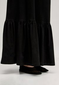Schwarzes Maxikleid mit vollem Saum und weicher Textur, kombiniert mit flachen schwarzen Schuhen mit glattem Finish und spitzem Fuß.
