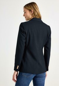 Marineblå skræddersyet blazer med et struktureret design, der har guldknapper på ærmerne og en glat tekstur. Båret over blå jeans.