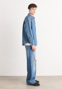 Camisa de denim y jeans desgastados en azul claro. La camisa presenta botones y un cuello clásico; los jeans tienen desgarros y un corte relajado. Zapatos negros.