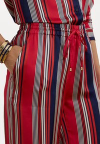 Red and navy striped trousers with an elastic waistband, drawstring detail, and side pockets; smooth fabric with metallic accents.