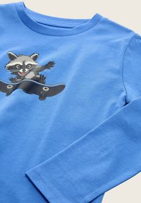 Maglietta blu a maniche lunghe in morbido cotone, con una grafica di un procione che skateboarda. Tessuto liscio con scollatura rotonda e polsini cuciti.