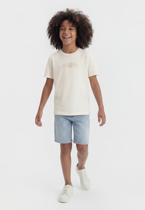 Copil zâmbitor cu păr creț care merge, purtând un tricou bej, pantaloni scurți de blugi albastru deschis și adidași albi, pe un fundal simplu.