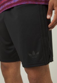 Fekete sportnadrág elasztikus derékkal, három függőleges csíkkal és egy diszkrét, sötét Adidas logóval az alsó bal oldalon.