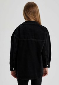 Chaqueta de mezclilla negra con un corte relajado, que presenta un puño abotonado y una costura en el yugo trasero, mostrada desde la parte posterior.