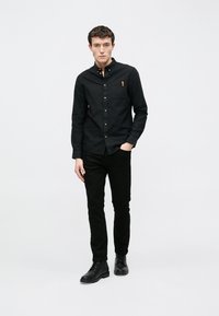 Cămașă neagră cu nasturi, cu buzunar la piept și etichetă maro, asortată cu pantaloni negri și bocanci negri cu șiret, pe un fundal alb simplu.