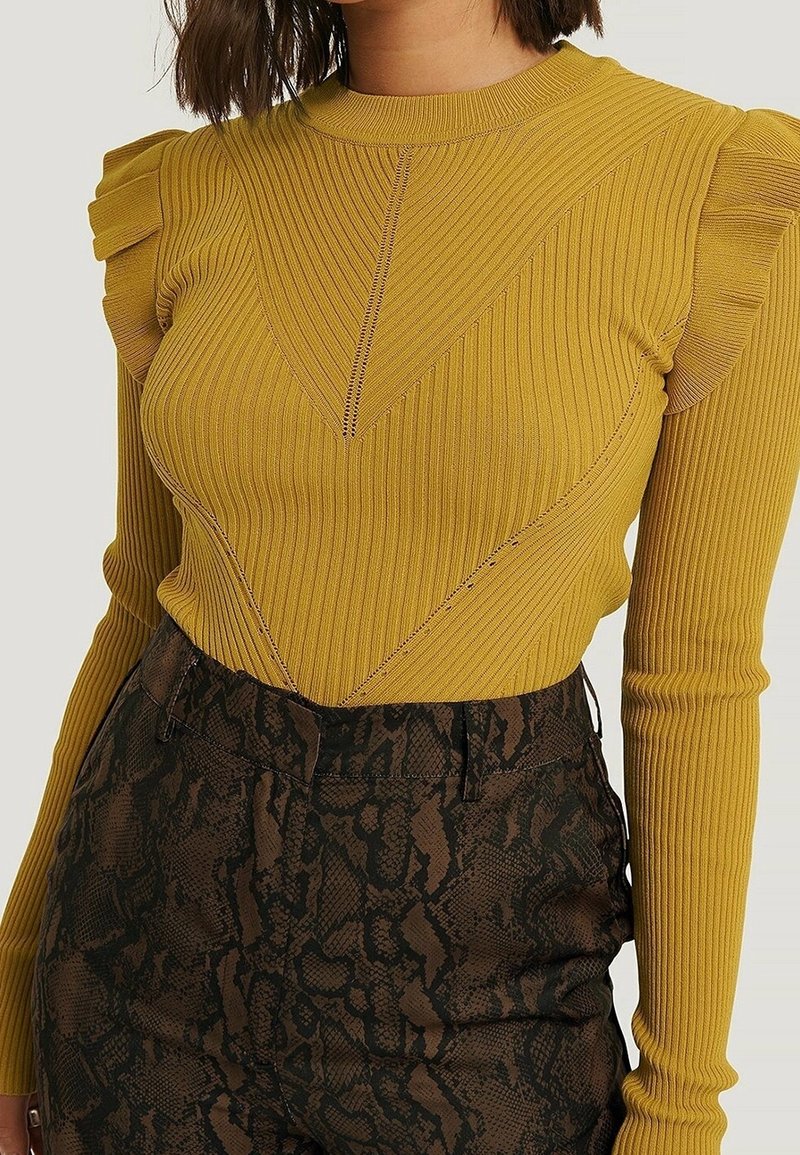 Pull en maille côtelée jaune moutarde avec des détails à volants aux épaules, associé à un pantalon taille haute marron foncé imprimé serpent.