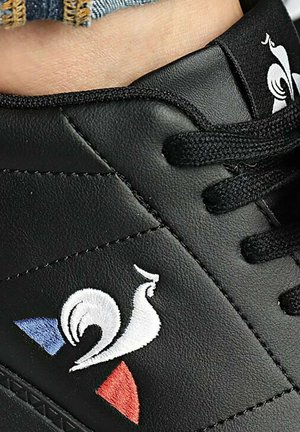 Black synthetic sneaker featuring a white embroidered rooster logo and red and blue accents on a textured side panel. Black laces.