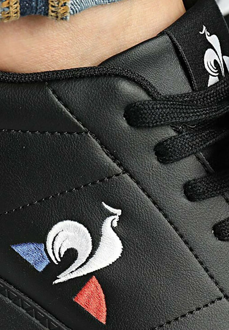 Baskets synthétiques noires avec un logo de coq brodé blanc et des accents rouges et bleus sur un panneau latéral texturé. Lacets noirs.