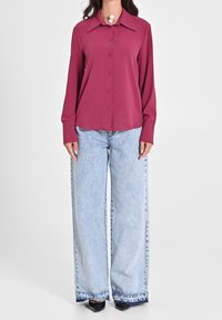 Femme portant un chemisier magenta à manches longues avec des boutons, un jean large bleu clair, des chaussures noires à bout pointu et un grand collier en perles blanches.