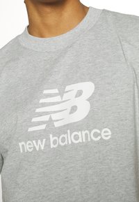 Primer plano de una persona que lleva una camiseta gris claro con un logo y texto en blanco de New Balance en el pecho.