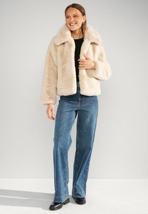 Donna sorridente che indossa una giacca in ecopelliccia beige, un top nero, jeans blu a gamba dritta e scarpe nere su uno sfondo grigio chiaro uniforme.