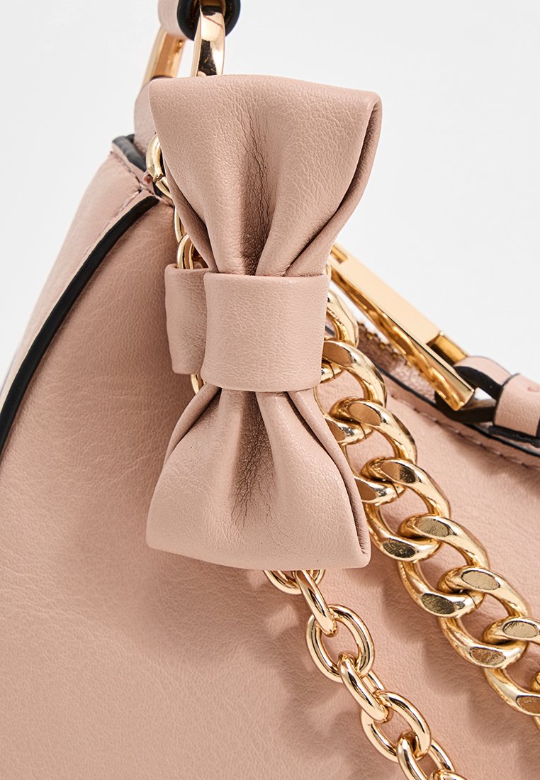 Gros plan sur un sac à main en cuir beige avec un nœud décoratif et une bandoulière en chaîne dorée sur un fond neutre.
