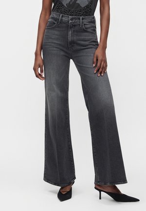Person trägt hoch taillierte, weit geschnittene ausgewaschene schwarze Jeans und schwarze spitze Schuhe, steht vor einem schlichten hellen Hintergrund.