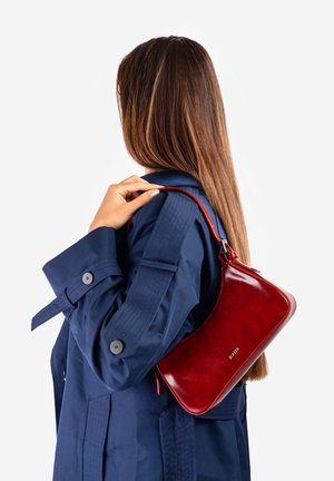 Femme aux longs cheveux bruns en manteau bleu marine tenant un petit sac à main en cuir rouge brillant à l'épaule sur fond blanc.