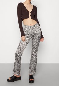 Brązowy sweter o skróconym kroju z długimi rękawami, z wycięciem z przodu i detalem w postaci pierścienia. Srebrne spodnie w wężowy wzór z dzwonami i gładką teksturą. Czarne sandały na platformie.