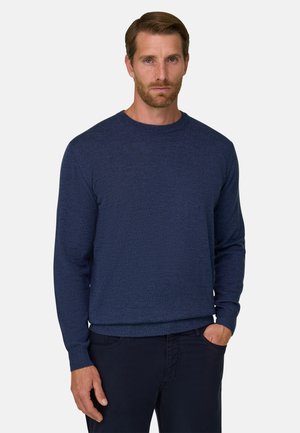 Homme aux cheveux courts bruns et à la barbe, portant un pull bleu foncé à manches longues et un pantalon noir, debout avec une main dans la poche.