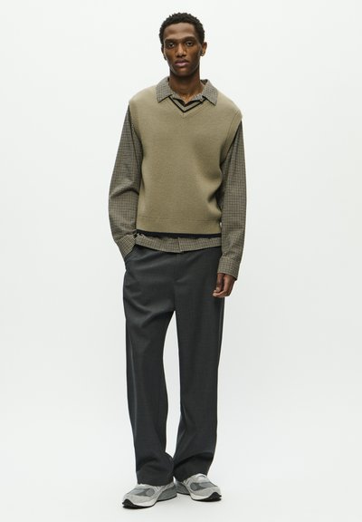 Gilet lavorato a maglia di un verde oliva chiaro sopra una camicia a motivi e quadri; pantaloni grigi su misura; sneakers grigie con accenti discreti.