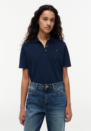 Frau mit langen lockigen Haaren, die ein dunkelblaues Poloshirt mit kurzen Ärmeln und blaue hoch taillierte Jeans trägt, steht vor einem schlichten Hintergrund.