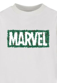 Valkoinen t-paita, jossa on sana "MARVEL" paksuilla valkoisilla kirjaimilla, ympäröitynä vihreällä kasvillisuudella ja punaisilla yksityiskohdilla.
